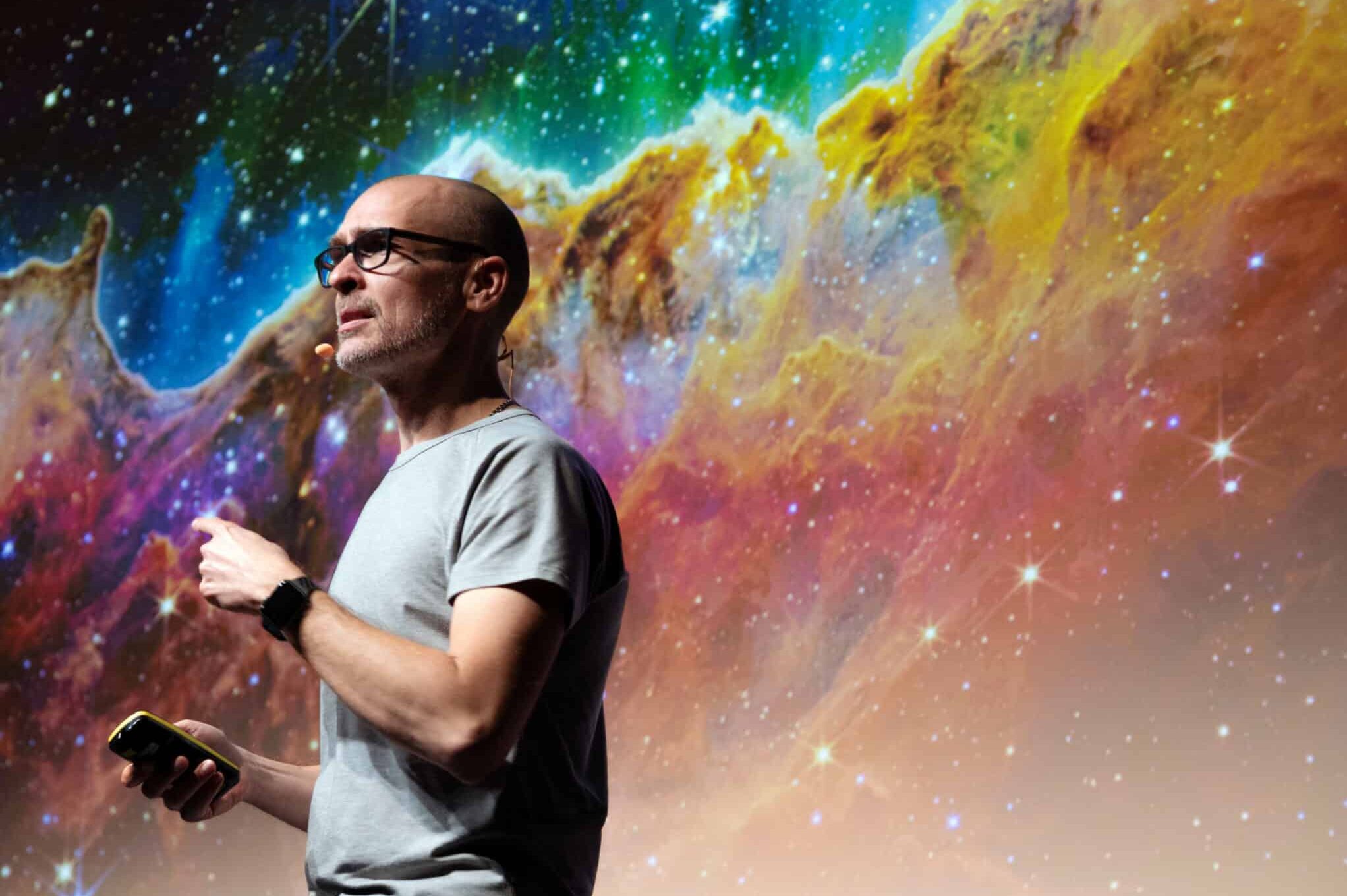 Matteo Cassese on stage presenting behind a space themed slide with colorful cosmic nebula background.